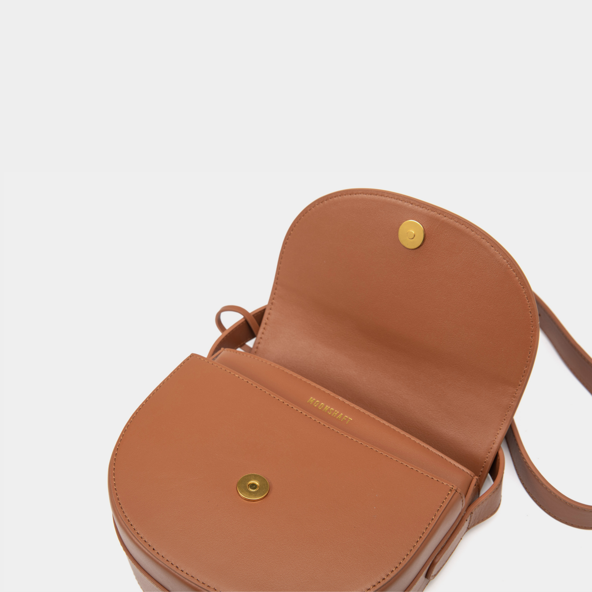 The close-up view displays the interior of the caramel half-moon crossbody bag with its smooth leather flap open. A gold-tone magnetic closure is prominently featured, offering a secure and stylish fastening. The inner lining is minimalistic, with the brand name "MOONSHAFT" subtly embossed in gold just beneath the closure for a refined touch.