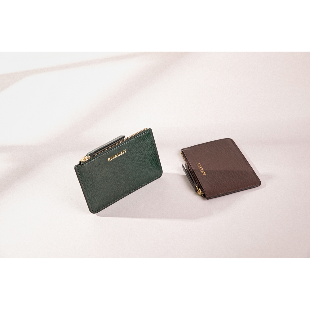 Featuring two leather cardholders placed on a light surface. The cardholder on the left is a dark green leather cardholder standing upright with the MOONSHAFT logo embossed in gold and a zipper closure on top. The cardholder on the right is a brown leather cardholder lying flat with the MOONSHAFT logo also embossed in gold. Both cardholders have gold-tone zipper hardware.