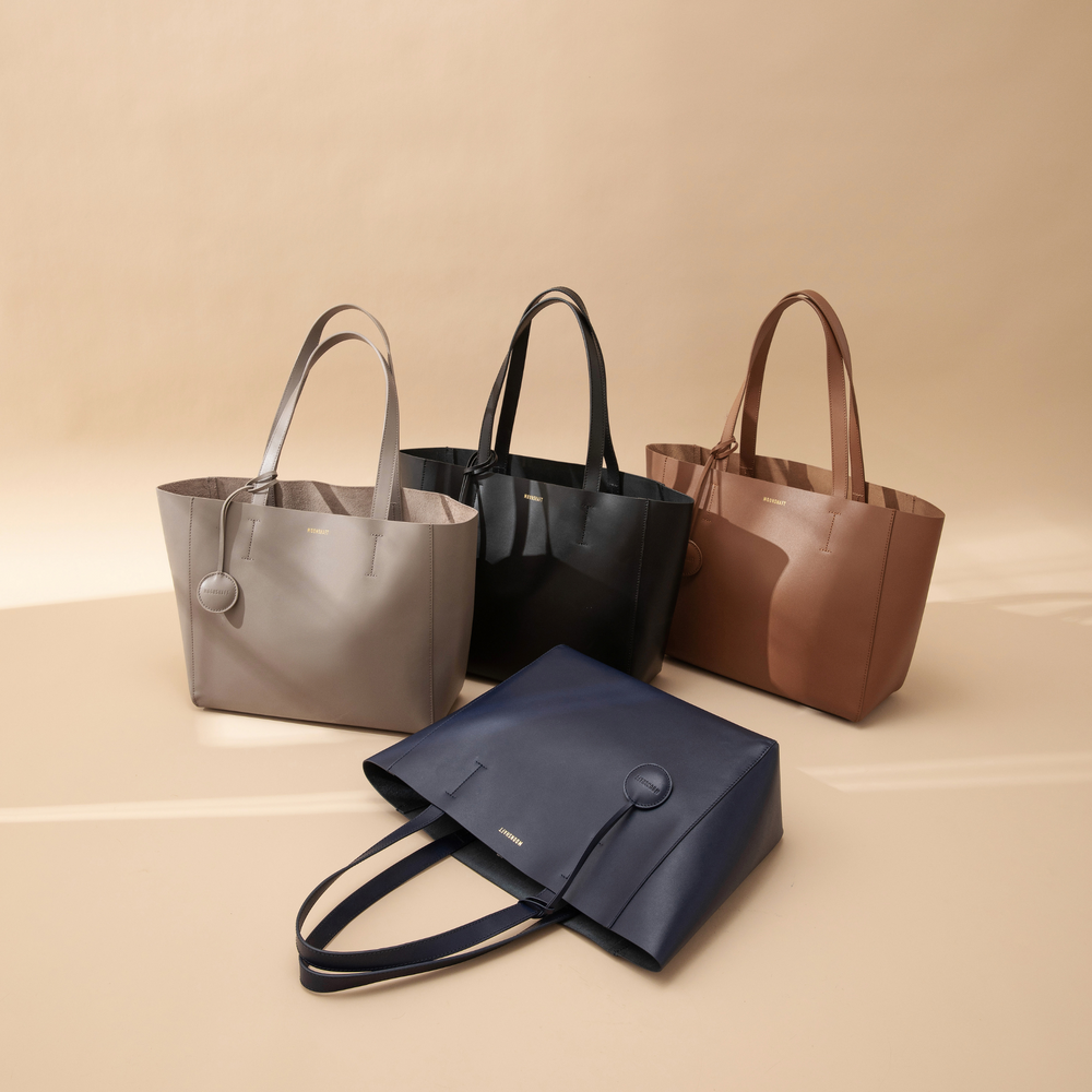 Four leather tote bags displayed in a creative arrangement, showcasing four different colors: taupe, caramel, black, and navy blue. Each bag features a minimalist, structured design with smooth leather surfaces and sturdy top handles. The bags are lined up side by side, highlighting the range of colors and the clean, elegant lines of each tote. The neutral and bold color options offer versatility for various outfits and occasions, making these bags both stylish and practical for everyday use.