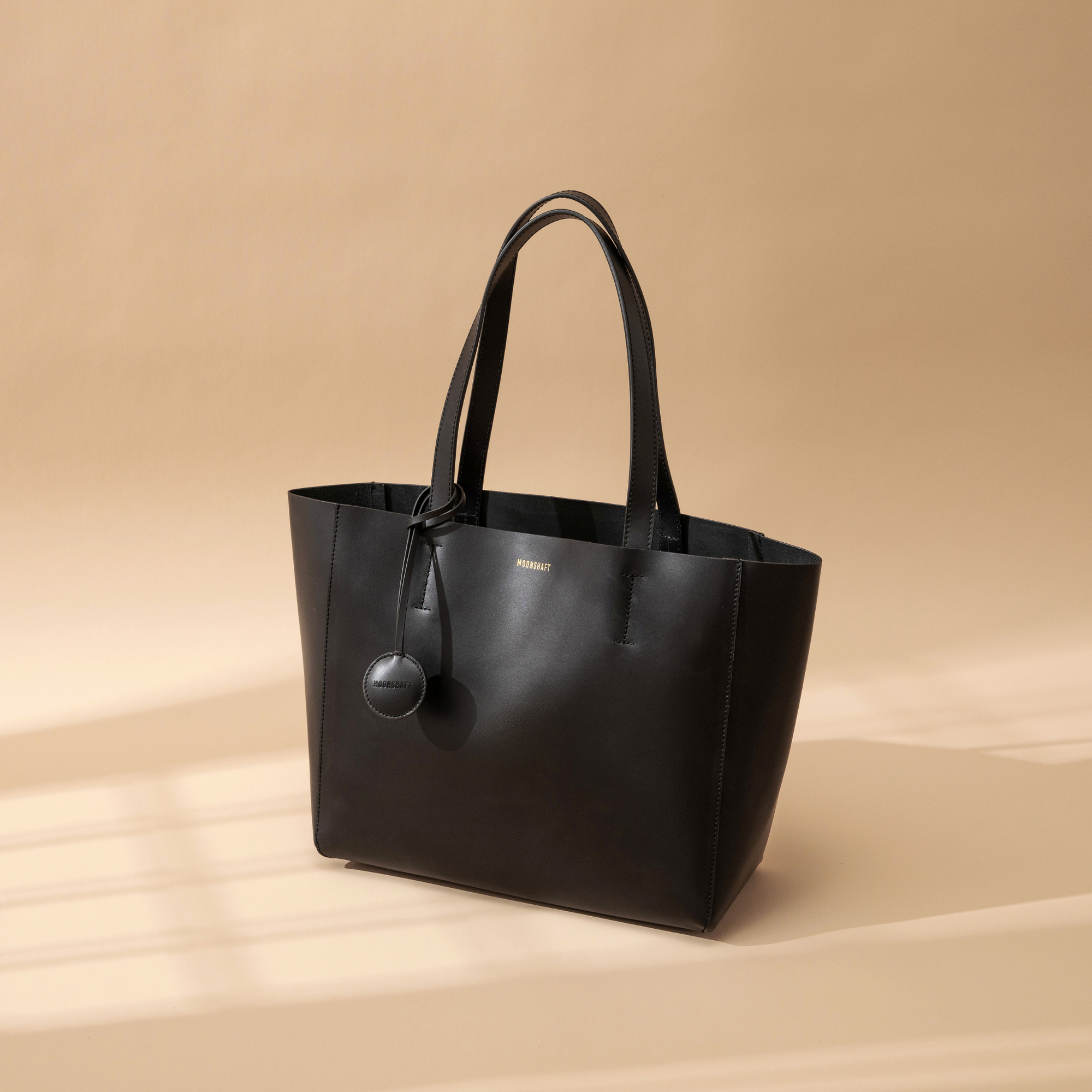 A black leather tote bag displayed at a creative diagonal angle against a plain background. The smooth leather surface and structured design are highlighted, emphasizing the bag's minimalist and elegant look. The tote features two sturdy leather handles and clean, sleek lines with no external embellishments. The neutral taupe color enhances its versatility, making it suitable for various styles and occasions. The diagonal orientation adds a dynamic and artistic presentation, showcasing the tote’s craftsmans