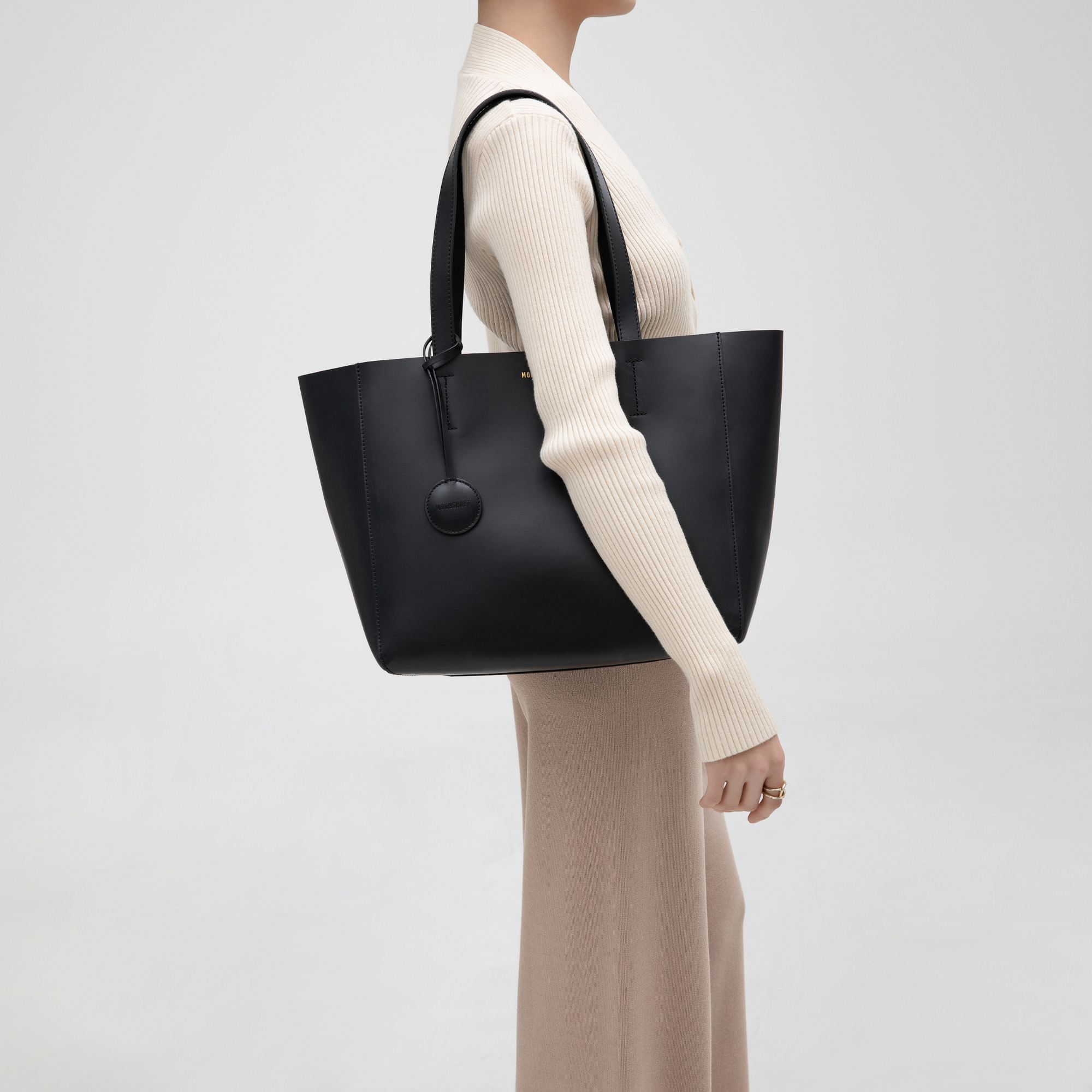A black leather tote bag modeled from the front, worn over the shoulder, showcasing its minimalist and structured design. The smooth leather surface and clean lines emphasize the tote’s elegant and sleek appearance. The long leather handles rest comfortably on the model’s shoulder, demonstrating its functionality. The black color adds a timeless and versatile appeal, making it suitable for both casual and professional settings. The tote’s spacious size makes it ideal for everyday use.