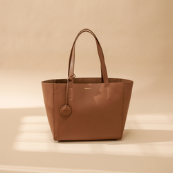 A caram leather tote bag displayed at a creative diagonal angle against a plain background. The smooth leather surface and structured design are highlighted, emphasizing the bag's minimalist and elegant look. The tote features two sturdy leather handles and clean, sleek lines with no external embellishments. The neutral taupe color enhances its versatility, making it suitable for various styles and occasions. The diagonal orientation adds a dynamic and artistic presentation, showcasing the tote’s craftsmans