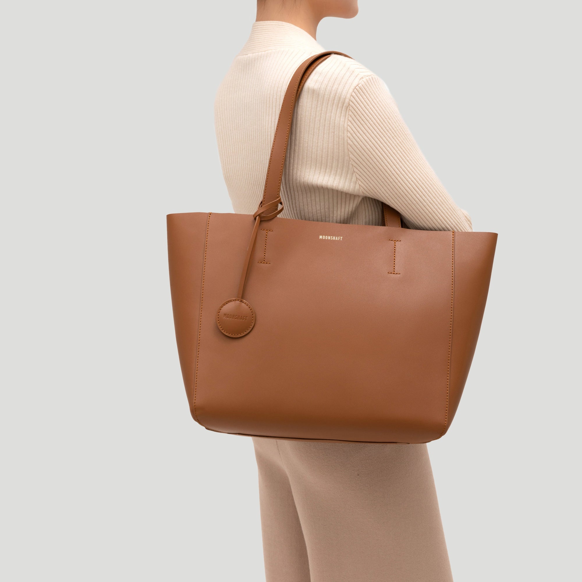 A caramel-colored leather tote bag modeled from the front, showcasing its minimalist and structured design. The smooth leather surface and clean lines emphasize the bag’s elegance, with two sturdy handles visible, allowing the bag to be comfortably held in hand. The rich caramel color adds warmth and versatility, making it suitable for a variety of outfits and occasions. The tote’s spacious size is ideal for carrying everyday essentials, blending style with practicality.