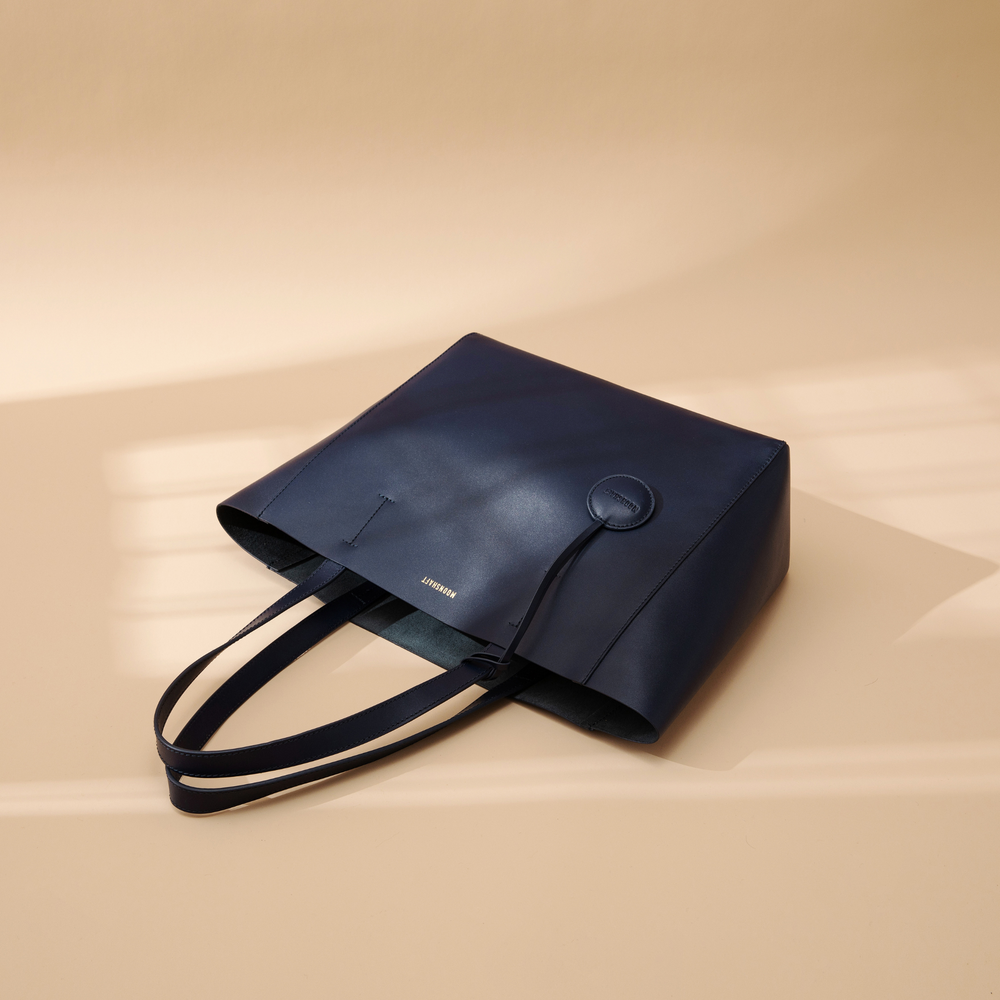 A navy leather tote bag displayed from a top-down view at a creative angle. The smooth leather surface and clean lines emphasize the bag's minimalist and structured design. The two sturdy leather handles are visible, and the spacious, open top highlights the tote's practicality for carrying everyday essentials. The rich navy color adds a sophisticated and versatile touch, making the bag suitable for various outfits and occasions while maintaining its sleek, modern aesthetic.