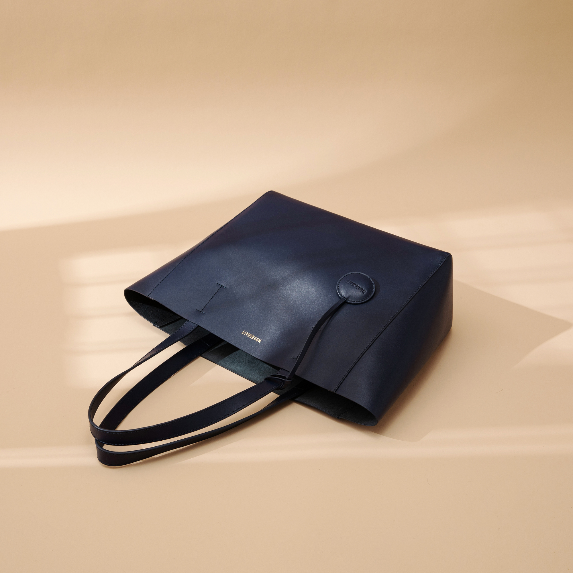 A navy leather tote bag displayed from a top-down view at a creative angle. The smooth leather surface and clean lines emphasize the bag's minimalist and structured design. The two sturdy leather handles are visible, and the spacious, open top highlights the tote's practicality for carrying everyday essentials. The rich navy color adds a sophisticated and versatile touch, making the bag suitable for various outfits and occasions while maintaining its sleek, modern aesthetic.