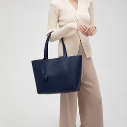 A navy blue leather tote bag modeled by a person, highlighting its minimalist design and practicality. The bag is held in hand, showcasing its structured shape and smooth leather texture. The neutral taupe color complements the model's outfit, demonstrating the tote’s versatility for both casual and professional looks. The long leather handles and spacious size are clearly visible, making it ideal for carrying everyday essentials while maintaining a stylish, elegant appearance.