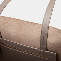 A close-up view of the interior of a taupe leather tote bag, showcasing its spacious and unlined compartment. The smooth leather exterior is visible along the edges, maintaining the minimalist design. The open top reveals ample room for carrying various items, emphasizing the tote's practicality. The sturdy leather handles are also partially visible, indicating the bag’s durability. The neutral taupe color adds a versatile and stylish appeal to the overall look.