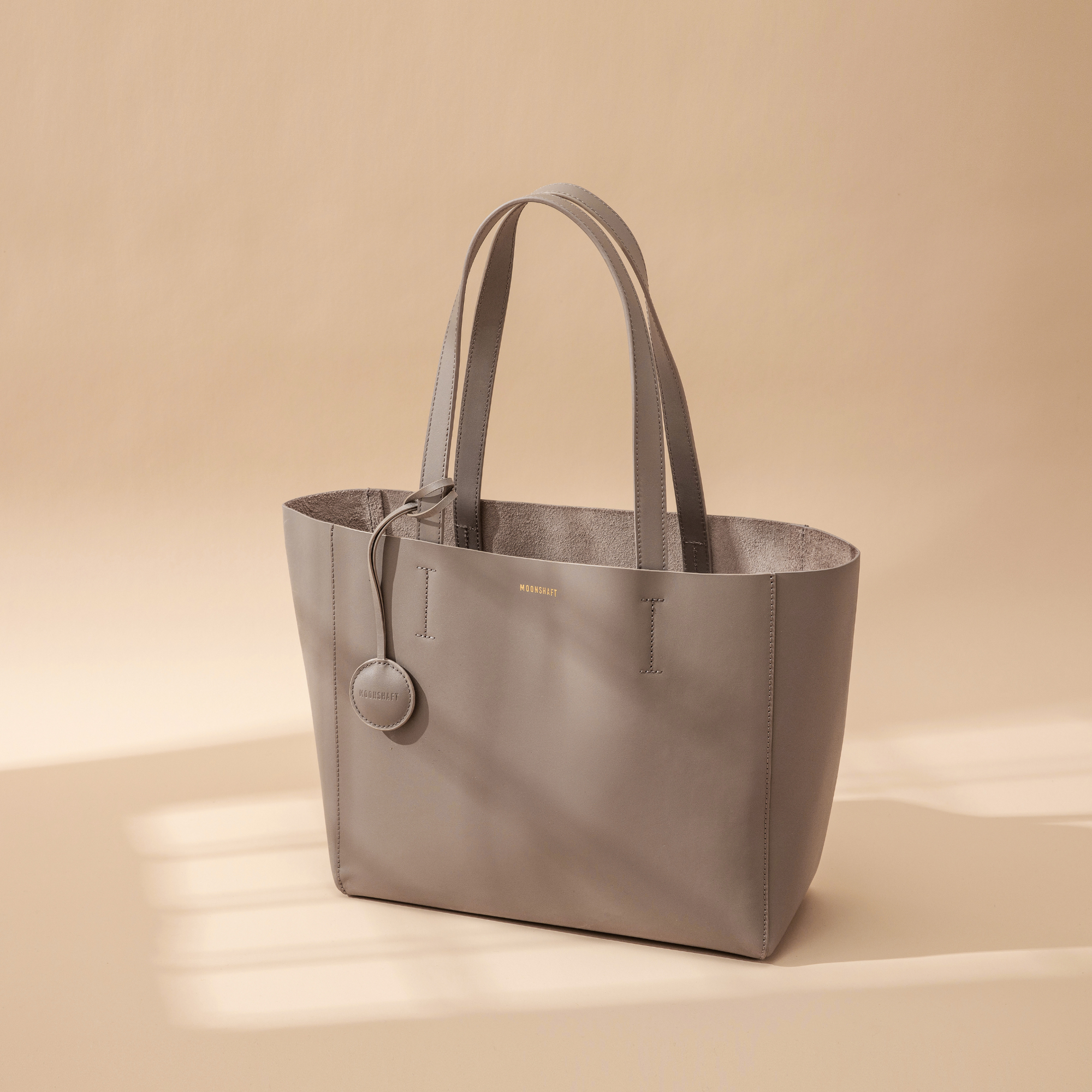 A taupe leather tote bag displayed at a creative diagonal angle against a plain background. The smooth leather surface and structured design are highlighted, emphasizing the bag's minimalist and elegant look. The tote features two sturdy leather handles and clean, sleek lines with no external embellishments. The neutral taupe color enhances its versatility, making it suitable for various styles and occasions. The diagonal orientation adds a dynamic and artistic presentation, showcasing the tote’s craftsmans