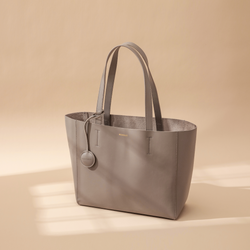 A taupe leather tote bag displayed at a creative diagonal angle against a plain background. The smooth leather surface and structured design are highlighted, emphasizing the bag's minimalist and elegant look. The tote features two sturdy leather handles and clean, sleek lines with no external embellishments. The neutral taupe color enhances its versatility, making it suitable for various styles and occasions. The diagonal orientation adds a dynamic and artistic presentation, showcasing the tote’s craftsmans