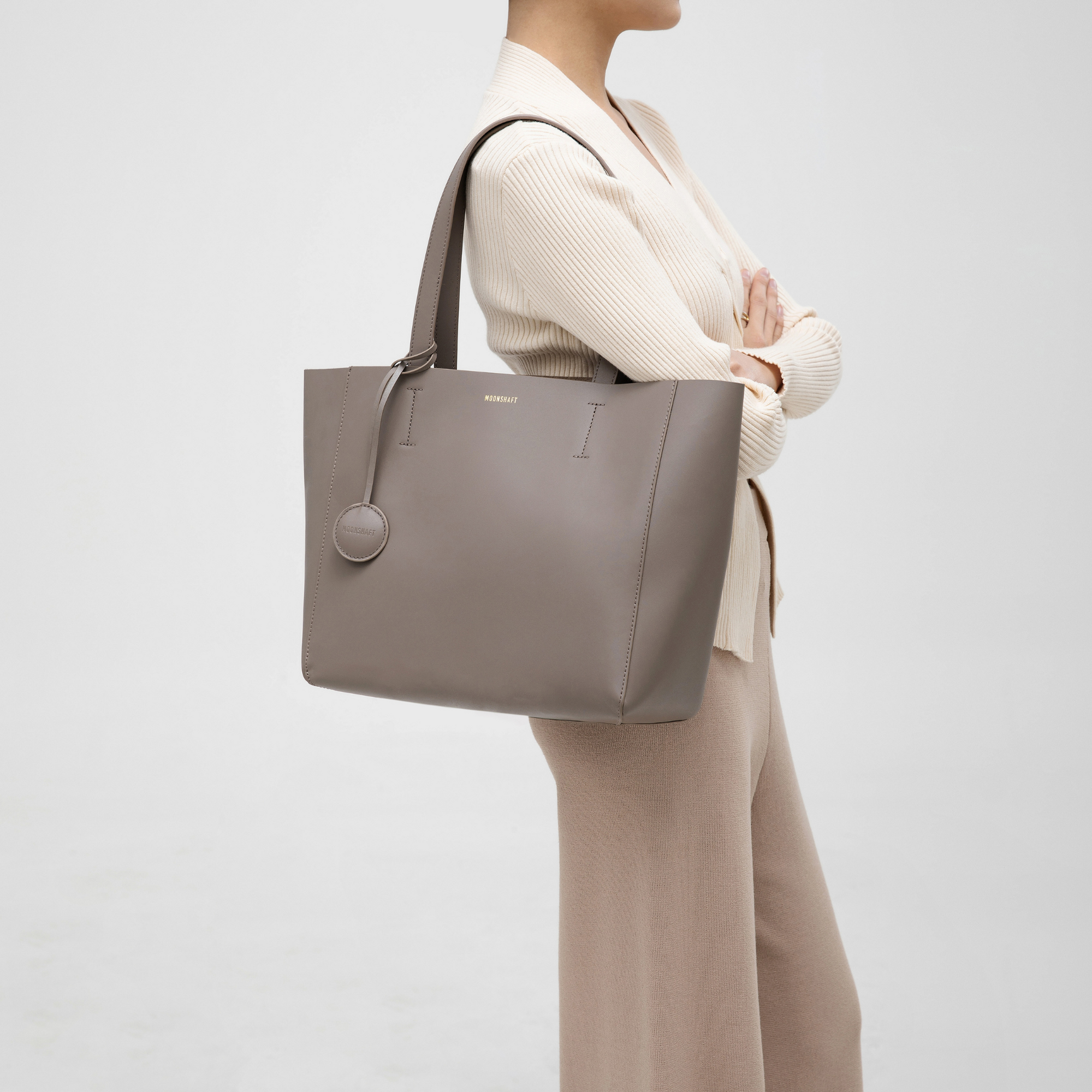 A taupe leather tote bag modeled on the shoulder, highlighting its minimalist and versatile design. The bag features a smooth leather texture and a structured, rectangular shape. The long handles rest comfortably on the model’s shoulder, demonstrating its ease of wear and functionality. The neutral taupe color complements the model's outfit, making the tote a stylish accessory for both casual and professional settings. The spacious design is ideal for carrying everyday essentials.