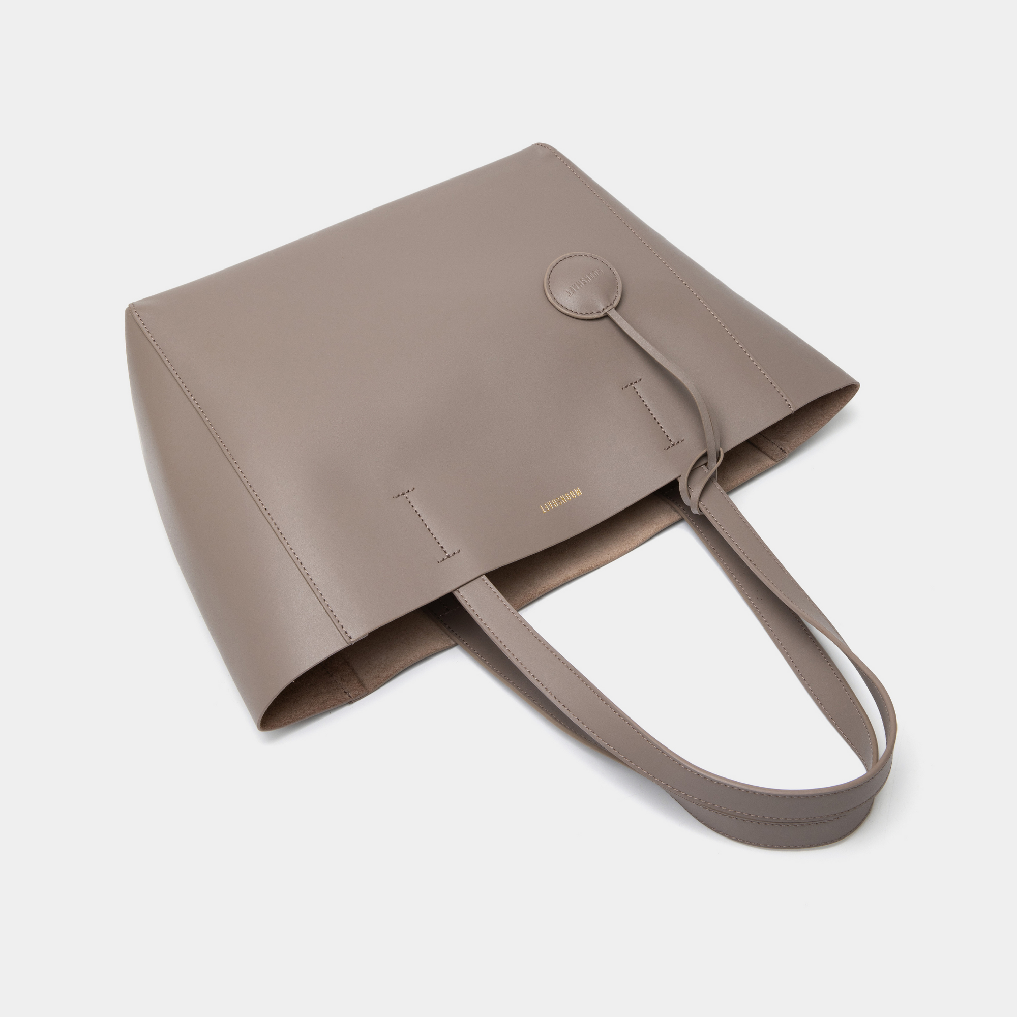 A taupe leather tote bag displayed from a top-down view with the bag closed. The minimalist design is highlighted by the smooth leather surface and clean lines. The two sturdy leather handles are neatly positioned, showcasing the tote's structured form. The neutral taupe color enhances its versatility, making it suitable for various outfits and occasions. The closed top view emphasizes the bag’s sleek and practical design, ideal for everyday use.